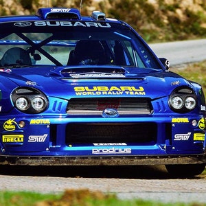 Op de afbeelding: Een blauwe Subaru World Rally Team auto met gele en zwarte accenten. De auto heeft een grote voorbumper en een Subaru-logo op de grille.