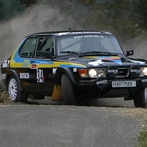 May include: A black Saab rally car with the number 34 on the side. The car is covered in dust and dirt, and the driver is wearing a helmet. The car has a "HARTMAN" sticker on the back.