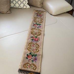 Könnte beinhalten: Ein langes, schmales, besticktes Blumentapete in Rosa-, Rot-, Lila- und Grüntönen. Die Tapete hat einen goldfarbenen Rand und eine Quaste am unteren Ende. Das Design zeigt sich wiederholende Blumenmuster.