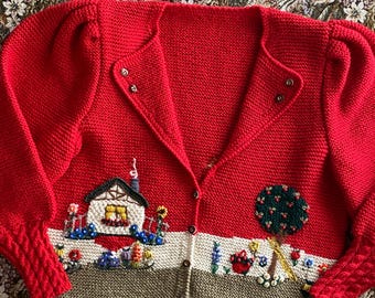 Op de afbeelding: Rood gebreid vest met een charmante geborduurde landschapsscène. De scène toont een huis, bloemen en een boom. Het vest heeft een knoopsluiting en getextureerde mouwen.