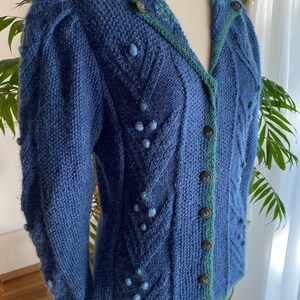 Può includere: Un cardigan blu in maglia con bordatura verde e chiusura con bottoni. Il cardigan presenta un motivo a treccia e  è decorato con piccole perline blu e verdi.