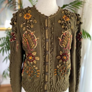 Può includere: Cardigan verde oliva con ricami floreali dettagliati in rosso, arancione e giallo. Il cardigan con bottoni presenta motivi a maglia strutturati e dettagli unici sulle spalle. Il design incorpora varie trame e colori.