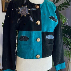 Può includere: Giacca in camoscio nera e verde acqua con applicazioni a tema celeste. La giacca presenta un design patchwork con dettagli di sole e nuvole, bottoni dorati a forma di stella e piccole borchie dorate. I polsini e l'orlo sono verde acqua.