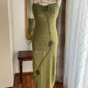 以下が含まれることがあります： オリーブグリーンのスパゲッティストラップドレス、フローラルアクセント付き。 ドレスは、シャーリングされたボディス、装飾的な花の装飾、そして流れるようなシアーなスカートが特徴です。 ドレスはマネキンに展示されています。