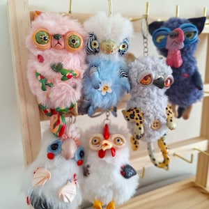 May include: Six colourful plush toys with unique features, including large eyes, patterned fur, and whimsical details. The toys are hanging from a wooden shelf.