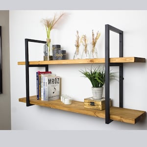 Op de afbeelding: Een zwarte metalen wandplank met twee houten planken. De planken zijn versierd met boeken, planten en decoratieve items. De plank is gemonteerd aan een witte muur.