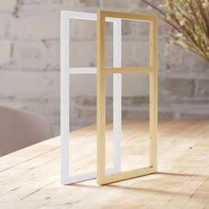 Può includere: Due cornici per foto, una bianca e una dorata, sono appoggiate su un tavolo di legno. Entrambe le cornici hanno un design semplice e rettangolare con un solo divisore orizzontale.