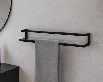 Puede incluir: Un toallero negro de doble barra montado en una pared de azulejos gris claro. Una toalla gris cuelga del toallero. El accesorio de baño tiene un diseño minimalista. Un grifo negro y un espejo redondo son visibles en el fondo.