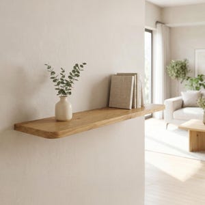 Pode incluir: Uma prateleira flutuante de madeira com acabamento natural é montada em uma parede de cor neutra. Um pequeno vaso branco com folhagem verde e vários livros são exibidos na prateleira. A prateleira tem um design simples e moderno.