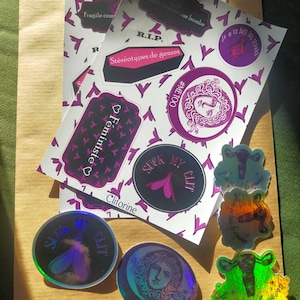 May include: A collection of stickers with various feminist themes. The stickers are printed on white paper with a pink and purple color scheme. The stickers feature phrases like "Feministe", "Clitorine", "Suck My Clit", and "#MeToo".