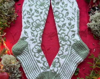 May include: A pair of knitted socks with a white floral pattern on a green background. The socks have green cuffs, heels, and toes. The socks are displayed on a red surface with decorative elements.