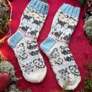 Peut inclure: Une paire de chaussettes tricotées sur le thème de l'hiver. Les chaussettes sont principalement blanches avec des poignets et des talons bleus. Elles présentent un motif de chats gris, de flocons de neige et d'autres formes géométriques.