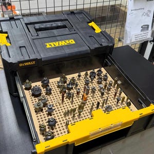 Op de afbeelding: Een zwarte DeWalt TSTAK opbergkoffer met een gele handgreep en sluiting, voorzien van een open lade met een beige geperforeerde organizer gevuld met diverse metalen freesbits in verschillende vormen en maten in een rasterpatroon.