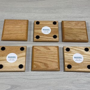 Puede incluir: Seis posavasos cuadrados de madera clara están dispuestos sobre una superficie gris claro. Cada posavasos tiene forma cuadrada con bordes redondeados. Algunos posavasos tienen una pegatina blanca con texto negro y almohadillas circulares negras.