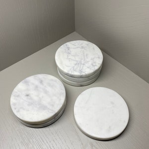 May include: Three white marble coasters with a veined pattern. The coasters are round and have a smooth surface.