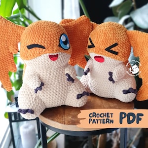 Könnte beinhalten: Zwei handgefertigte Häkeltier-Plüschtiere in Form einer Zeichentrickfigur, mit orangefarbenen und cremefarbenen Farben. Die Spielzeuge haben große orangefarbene Flügel, schwarze Akzente und blaue Augen. Der Text "CROCHET PATTERN PDF" ist sichtbar.