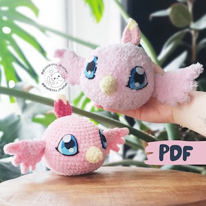 May include: Two pink crocheted bird plushies with blue eyes and yellow beaks. The plushies are sitting on a wooden surface. The image includes the text "PDF".