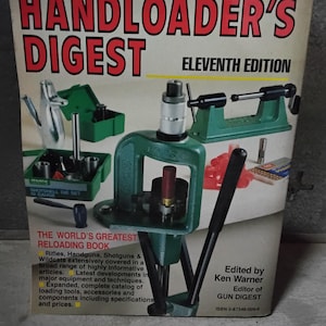 Puede incluir: La portada del libro "Handloader's Digest" Eleventh Edition presenta equipos y herramientas de recarga. El título del libro está en letras rojas grandes, con una prensa de recarga verde y otras herramientas. El libro está editado por Ken Warner.