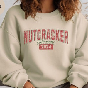 May include: A sand-colored sweatshirt with the text "NUTCRACKER Season 2024" in red and green lettering. The sweatshirt has a crew neck and long sleeves. The overall theme is holiday apparel.