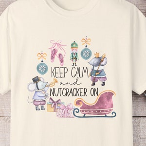 May include: Ivory-colored t-shirt with a whimsical Nutcracker-themed graphic. The design features ballet slippers, a toy soldier, and mice with musical instruments, along with the text "Keep Calm and Nutcracker On". The shirt also has the text "Comfort Colors".