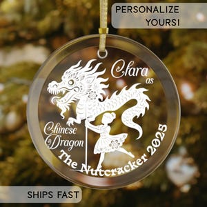 Puede incluir: Adorno navideño redondo y transparente con una cinta dorada. Presenta una ilustración blanca de un dragón chino y una niña, con el texto "Clara as Chinese Dragon The Nutcracker 2025".