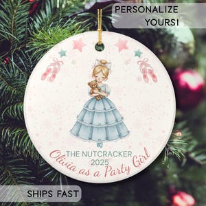 May include: A round white ceramic Christmas ornament featuring a central illustration of a girl in a blue dress holding a teddy bear, surrounded by pink ballet slippers and stars. The text reads THE NUTCRACKER 2025 Olivia as a Party Girl.