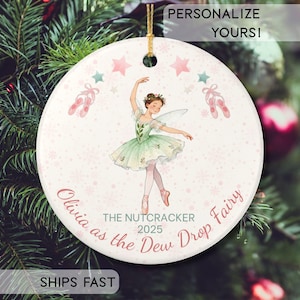Peut inclure: Une décoration de Noël ronde avec une ballerine en tutu vert et chaussons roses. L'ornement porte l'inscription "Olivia as the Dew Drop Fairy" et "The Nutcracker 2025". Le fond est rose clair avec des étoiles et des chaussons de ballet.