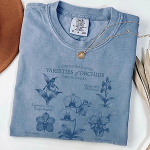 May include: A pale blue t-shirt with a botanical print of different orchid varieties. The shirt displays the text "A Concise Field Guide Varieties of Orchids Orchidaceae". A gold necklace and a brown hat are also visible.