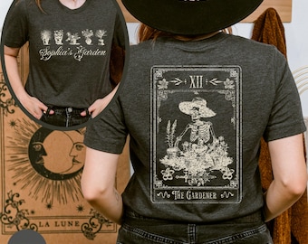 Puede incluir: Una camiseta gris oscuro con un gráfico blanco de una carta de tarot que representa un esqueleto haciendo jardinería. La carta está etiquetada como "The Gardener" y "XII". El texto "Sophia's Garden" está impreso en la parte delantera de la camiseta.