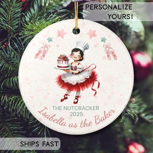 May include: A round white ceramic ornament featuring a graphic of a baker in a tutu holding a cake. The text reads "THE NUTCRACKER 2025" and "Isabella as the Baker". The design includes pink stars, ballet slippers, and snowflakes, finished with a gold hanging cord.