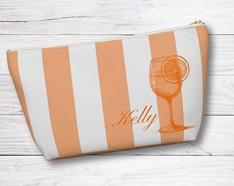 May include: A white and orange striped makeup bag with the name "Kelly" printed on it. The bag features a drawing of a glass of orange juice with a slice of orange.