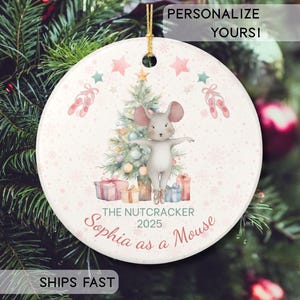 May include: Circular Christmas tree decoration with a mouse in ballet pose, in front of a Christmas tree. The ornament has the text "The Nutcracker 2025" and "Sophia as a Mouse". Pink stars and ballet shoes decorate the ornament.