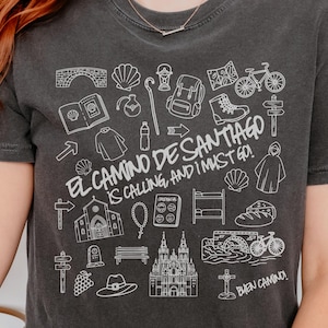 May include: Dark gray t-shirt with a white graphic design featuring travel-related illustrations and the text "EL CAMINO DE SANTIAGO IS CALLING, AND I MUST GO!" and "BUEN CAMINO!". The design includes a church, backpack, and bicycle.