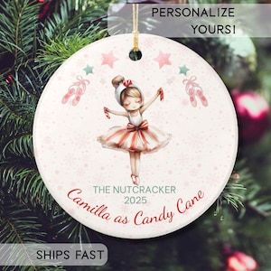 May include: A round white ceramic Christmas ornament featuring a ballerina illustration in a red and white striped tutu, holding candy canes. The text reads THE NUTCRACKER 2025 and Camilla as Candy Cane, surrounded by pink stars and ballet slippers.