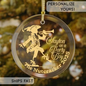 Peut inclure: Une décoration de Noël ronde et transparente avec un motif doré d'une personne jouant de la trompette. Le texte indique "Clayton as a Party Boy 2025" et "The Nutcracker". La décoration est suspendue à un ruban doré.