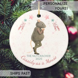 May include: A round Christmas ornament featuring a mouse character from The Nutcracker ballet. The ornament has a light pink background with snowflakes, stars, and ballet slippers. Text includes "The Nutcracker 2025" and "Cassidy as a Mouse".