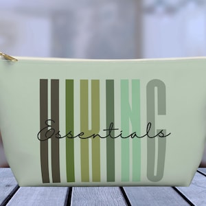 May include: A light green zippered pouch with a graphic design of vertical stripes in shades of green and brown. The text "Hiking Essentials" is written in black cursive font.