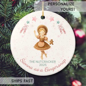 May include: A round white ceramic ornament featuring a central illustration of a child dressed in a brown gingerbread-themed outfit holding a cookie. The design includes pink ballet slippers, stars, and snowflakes. Text reads: THE NUTCRACKER 2025 Sienna as a Gingersnap.