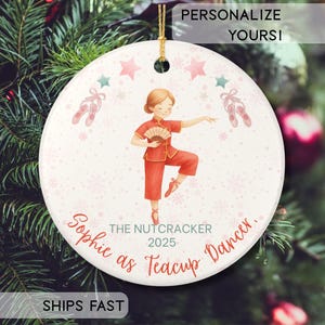 Puede incluir: Adorno navideño redondo con una bailarina con un traje rojo, sosteniendo un abanico. El adorno tiene el texto "Sophie as Teacup Dancer, The Nutcracker 2025". Estrellas rosas, zapatillas de ballet y copos de nieve decoran el fondo. El texto "Personalize Yours!" está en la parte superior.
