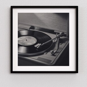 May include: Black and white photograph of a vintage record player with a vinyl record. The record player is in a wooden case, and the record is spinning. The image is framed in a black frame.