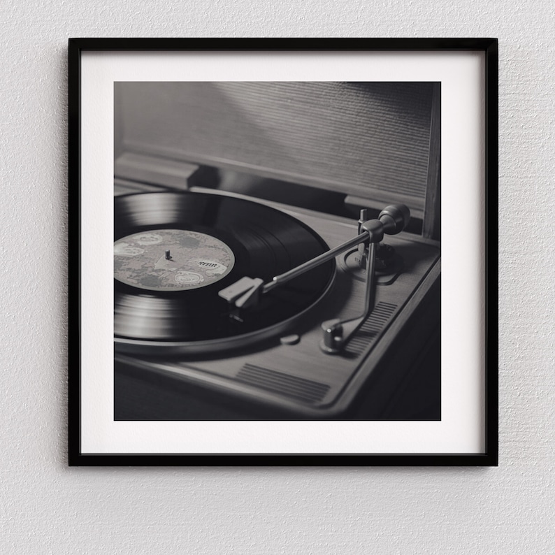 May include: Black and white photograph of a vintage record player with a vinyl record. The record player is in a wooden case, and the record is spinning. The image is framed in a black frame.