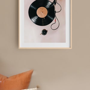 May include: A framed print of a black vinyl record on a turntable with a red needle. The record is on a light pink background.