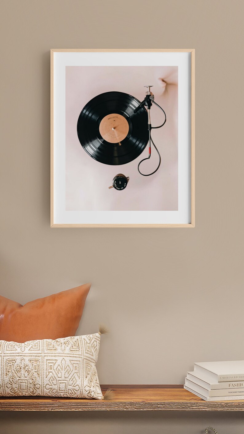 May include: A framed print of a black vinyl record on a turntable with a red needle. The record is on a light pink background.