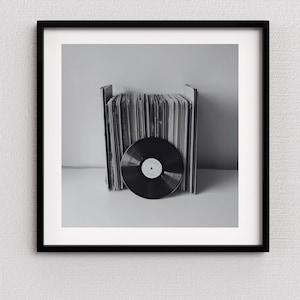 May include: Black and white framed print featuring a collection of vinyl records. A single record is in the foreground, with a stack of album covers behind it. The image is a monochromatic, minimalist composition.