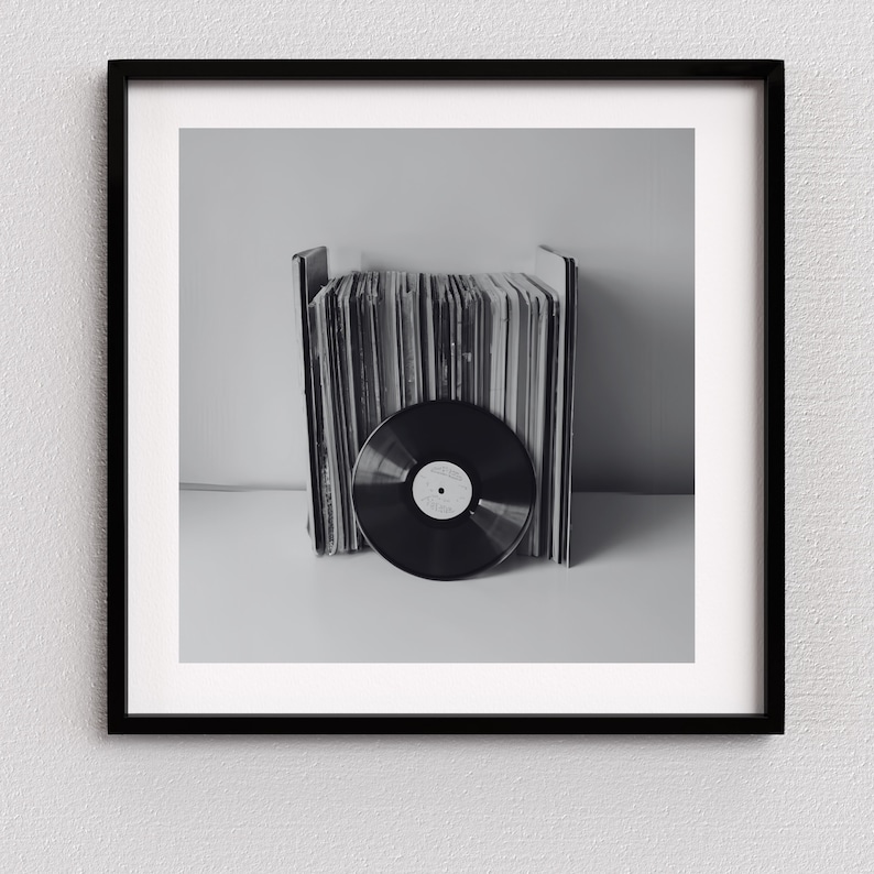 May include: Black and white framed print featuring a collection of vinyl records. A single record is in the foreground, with a stack of album covers behind it. The image is a monochromatic, minimalist composition.