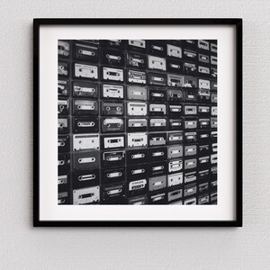 May include: Black and white framed print featuring a wall of vintage cassette tapes. The tapes are arranged in a grid pattern, showcasing a variety of labels and designs. The image is a nostalgic nod to the era of analog music.