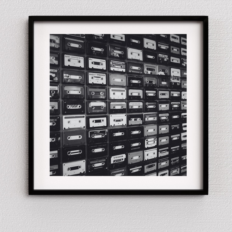 May include: Black and white framed print featuring a wall of vintage cassette tapes. The tapes are arranged in a grid pattern, showcasing a variety of labels and designs. The image is a nostalgic nod to the era of analog music.