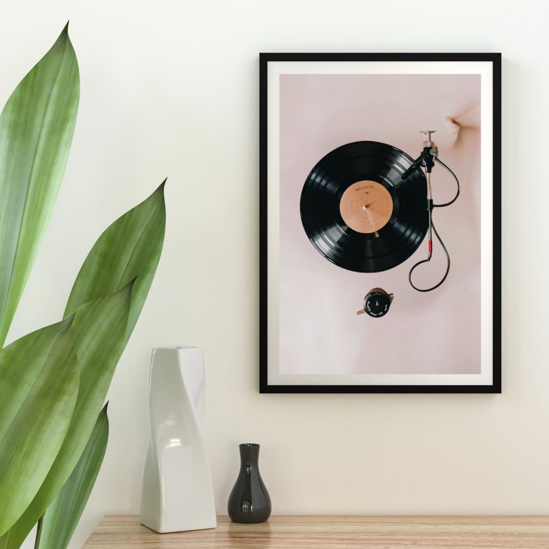May include: A framed black and white photograph of a black vinyl record on a turntable. The record is playing and the needle is visible. The background is a light pink.