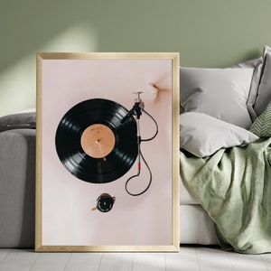 May include: A framed print of a vintage record player with a black vinyl record on the turntable. The record player is on a light pink background.