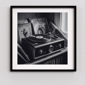 May include: Black and white photograph of a vintage record player with a vinyl record on the turntable. The record player is on a shelf with several vinyl records in the background. The image is framed in a black square frame.